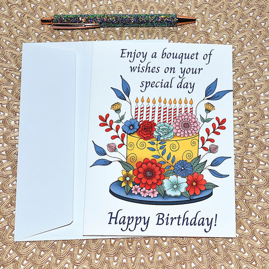 Birthday card with colorful cake illustration of flowers and candles with white envelope and glitter pen on a textured surface