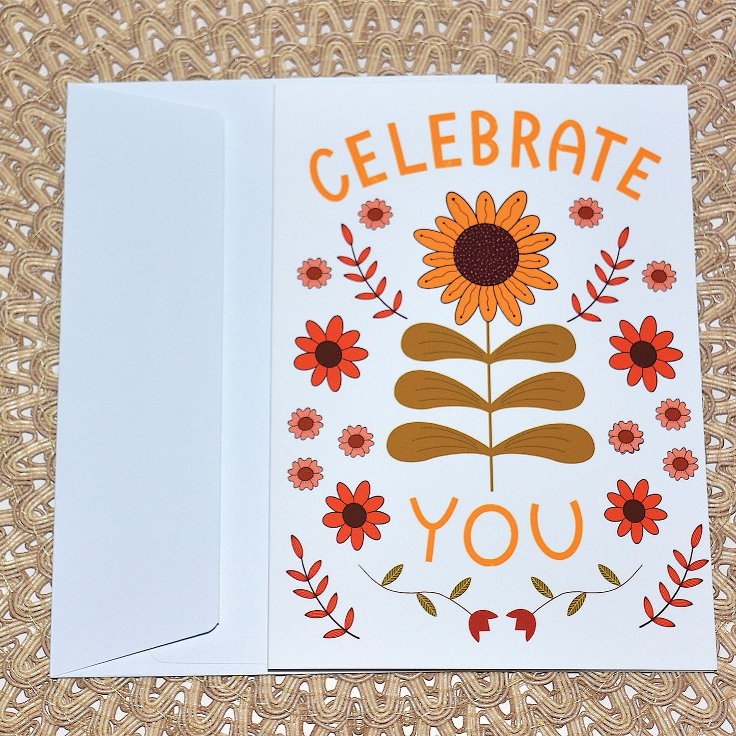 Greeting card with the words "celebrate you" adorned with sunflower leaves and flowers with white envelope on textured background