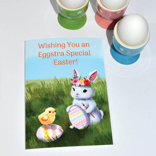 Easter card with bunny wearing floral headdress painting an easter egg and a chick standing on egg, in green pasture staged next to colorful egg cups