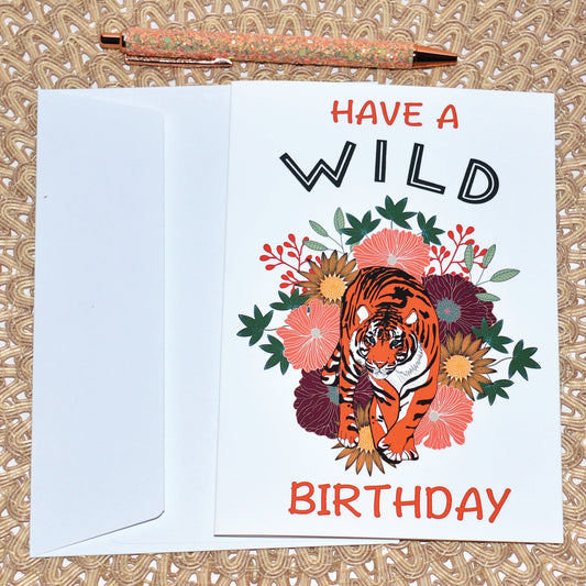 Greeting card with a tiger illustration and floral design, featuring the text 'Have a Wild Birthday' with white envelope and glitter pen on a textured surface.