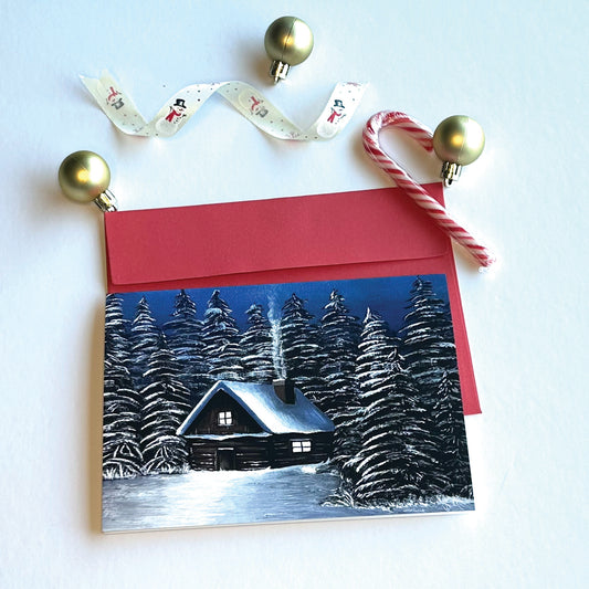 Christmas card with a snowy log cabin with evergreens landscape with red envelope and decorated with ornaments and ribbons.
