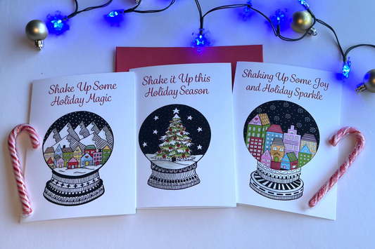 Three Christmas-themed greeting cards with snow globe designs on a white surface with candy canes and string lights.
