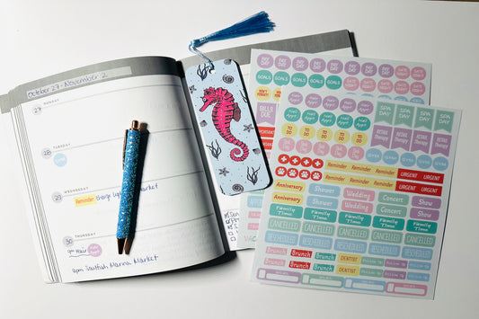 Open planner with planner sticker sheets, a seahorse bookmark and glitter pen on a white surface