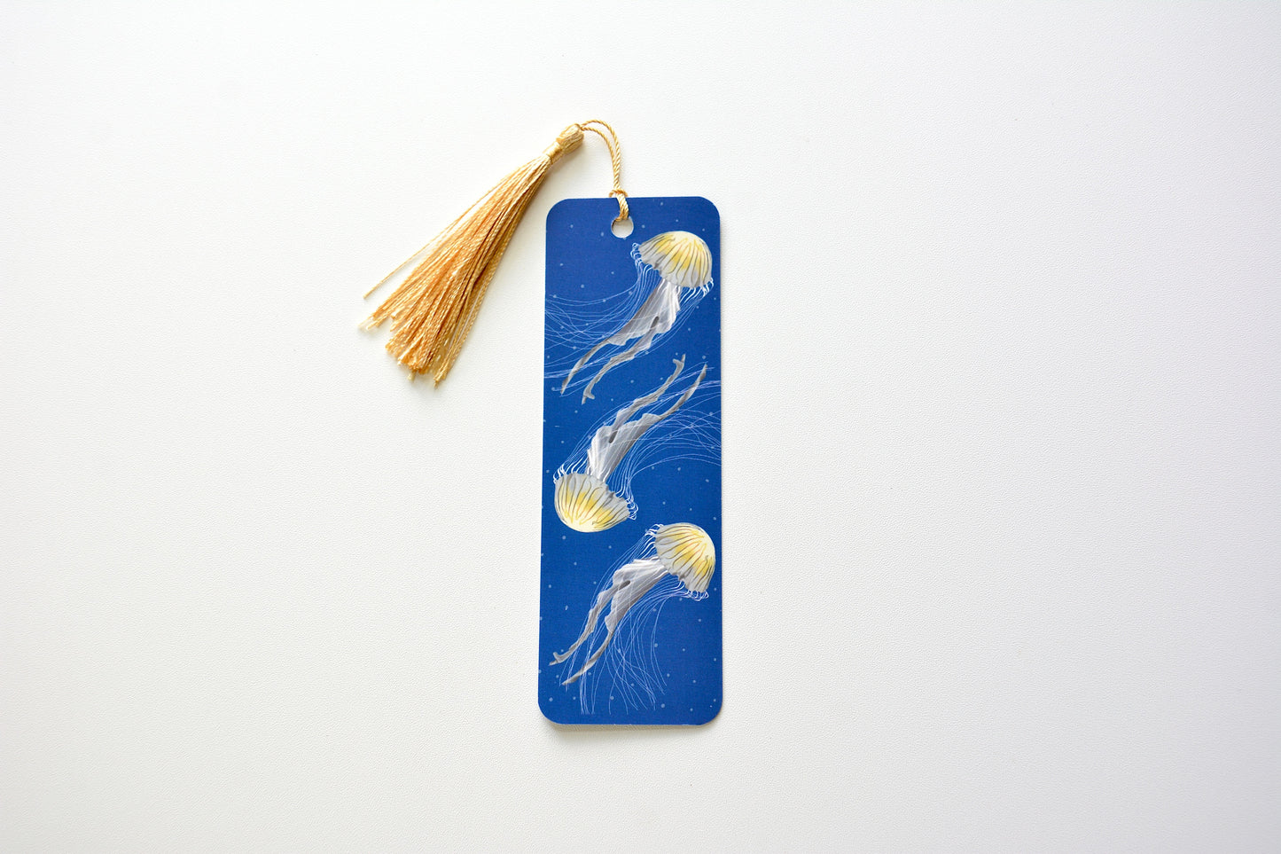 Bookmark with jellyfish design and gold tassel on a blue background placed on white surface