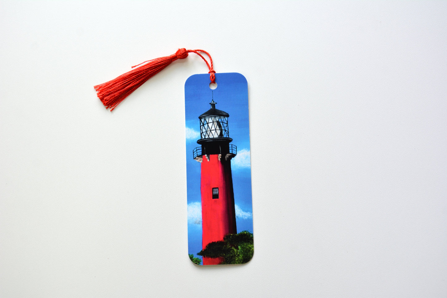 Bookmark with a Jupiter lighthouse design and red tassel  with clouds and sky background placed on a white surface
