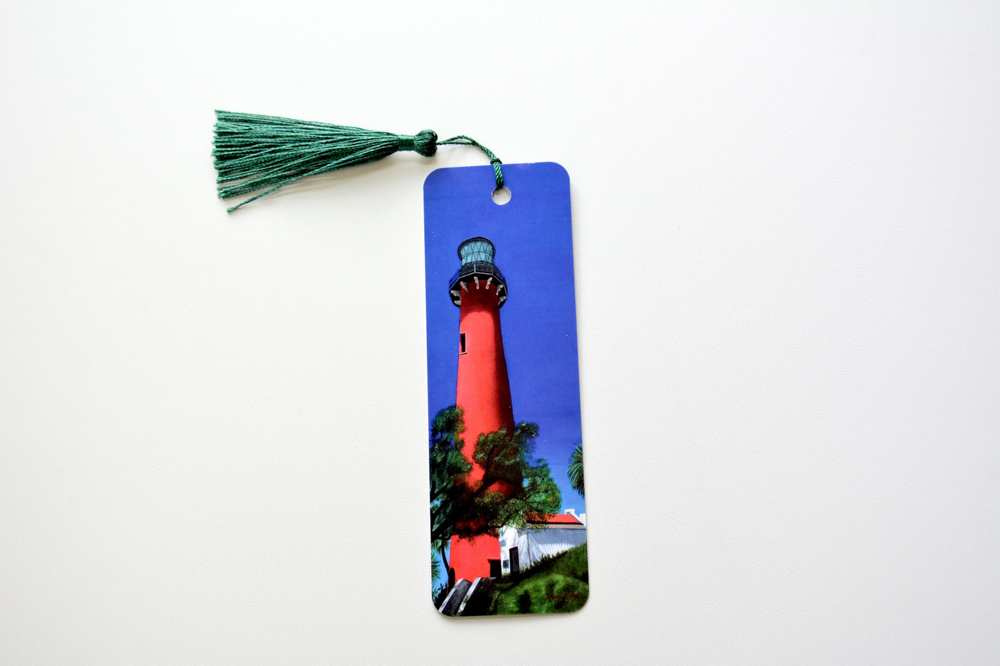 Bookmark with the Jupiter Lighthouse design with white house and trees on a blue background and green tassel placed onto a white surface