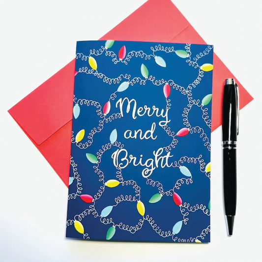 Blue card with Christmas lights and 'Merry and Bright' text, red envelope, and black pen on a white background