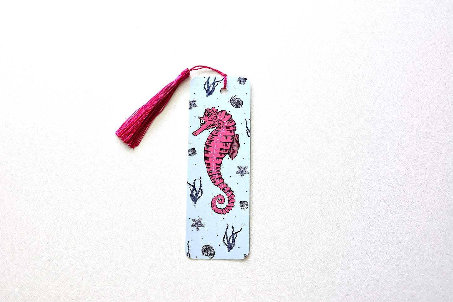 Bookmark with pink seahorse and a background of a pattern with sea kelp, starfish and seashells on a light blue background with pink tassel placed onto a white surface