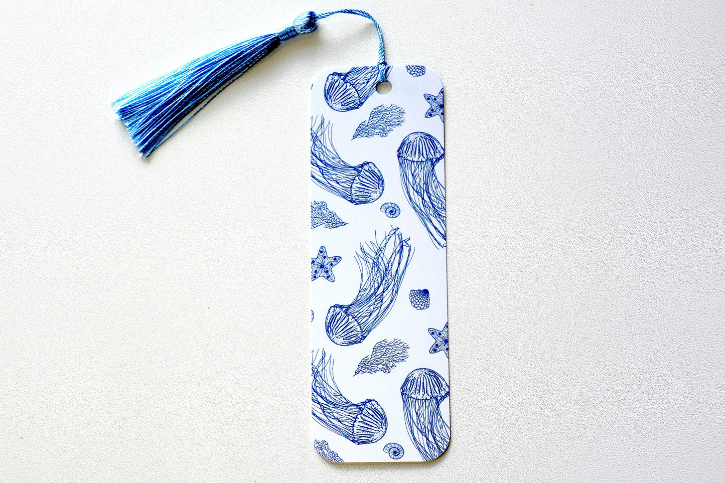 Bookmark with navy blue sea-themed pattern with sea kelp, jellyfish, seashells and starfish on light blue background with a blue tassel placed on a white surface