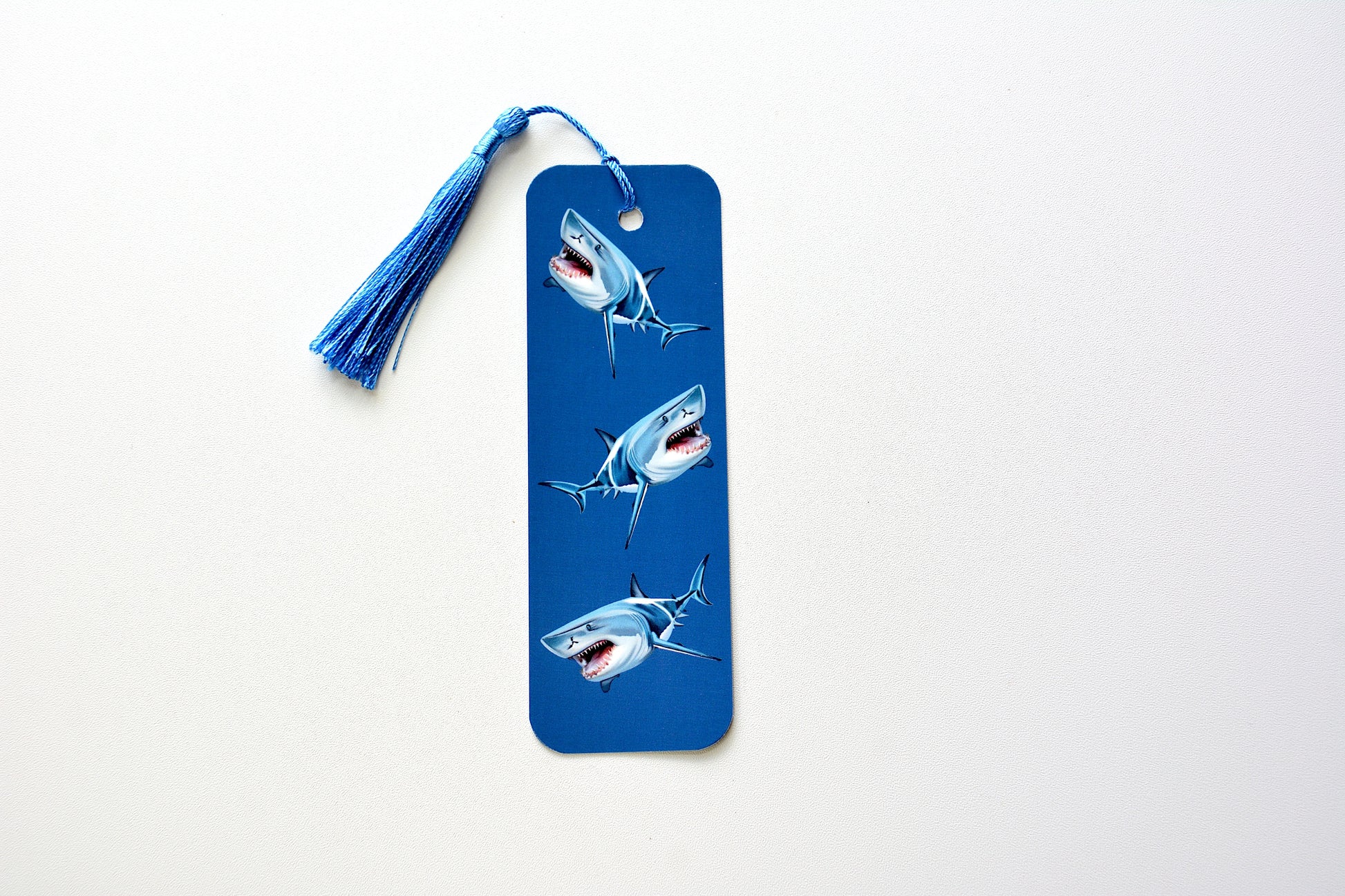 Bookmark with shark illustrations and a blue tassel on a blue background placed on a white surface