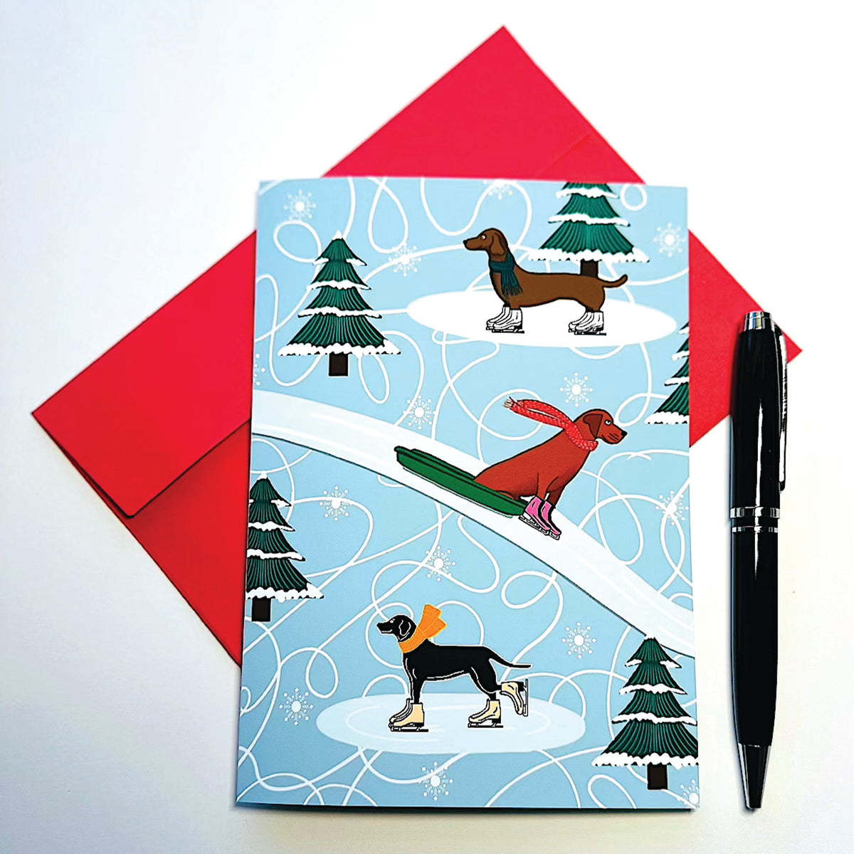 Holiday card with winter-themed illustration of dogs ice skating and on sled with scarves and a red envelope and pen on a white background.